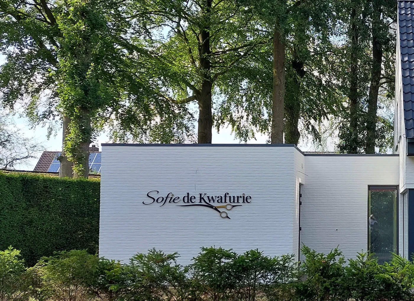 De witte gevel van Sofie de Kwafurie met het logo, tussen hoge bomen