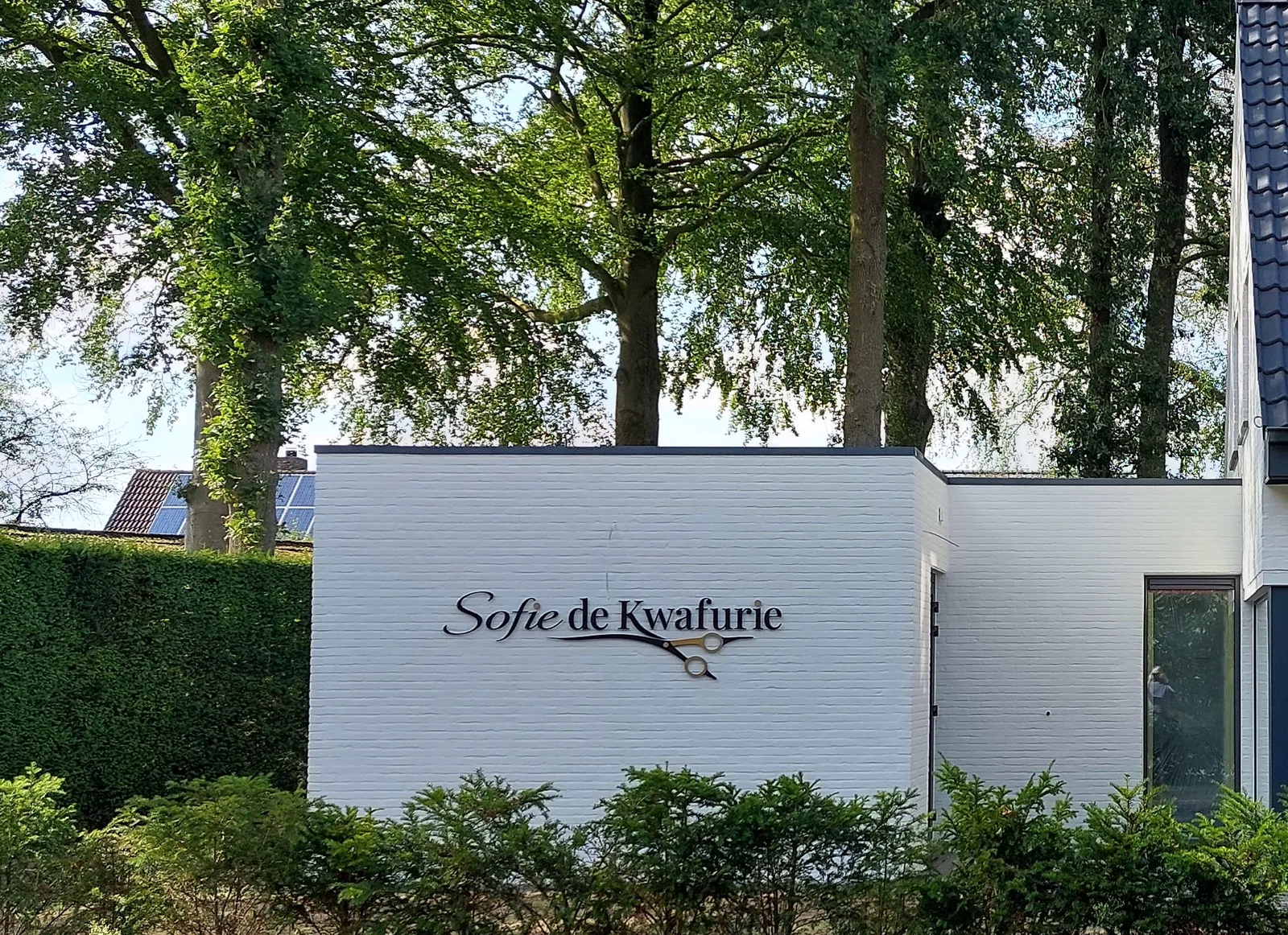 De witte gevel van Sofie de Kwafurie met het logo, onder de hoge bomen naast het salon