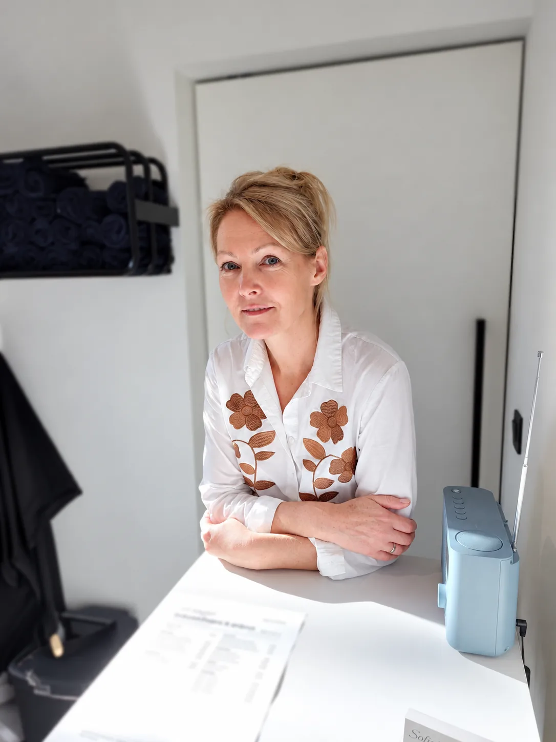 Sofie aan de toog in het salon, met opgerolde handdoeken op het rek achter haar